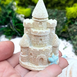 Könnte beinhalten: Eine kleine, beige, texturierte Schlossfigur mit einem kegelförmigen Dach, Zinnentürmen und Bogenfenstern. Die Oberfläche weist ein dezentes Glitzer-Finish auf und ist an der Basis mit einem hellblauen Seestern-Akzent versehen.
