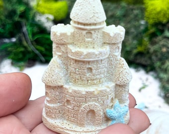 Könnte beinhalten: Eine kleine, beige, texturierte Schlossfigur mit einem kegelförmigen Dach, Zinnentürmen und Bogenfenstern. Die Oberfläche weist ein dezentes Glitzer-Finish auf und ist an der Basis mit einem hellblauen Seestern-Akzent versehen.