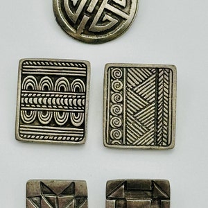 Könnte beinhalten: Fünf silberfarbene Metallknöpfe mit verschiedenen geometrischen Mustern. Die Knöpfe sind quadratisch und rund.