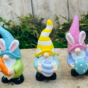 May include: Three Easter gnomes with bunny ears. The gnome on the left is holding a carrot, the gnome in the middle is holding a striped egg, and the gnome on the right is holding a blue and white speckled egg.
