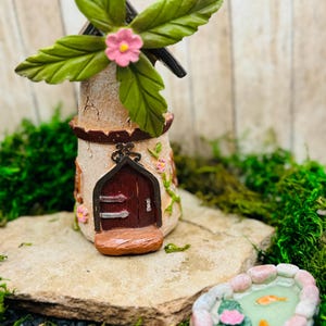 May include: A miniature fairy garden windmill with a pink flower on top, a brown door, and a stone base. A small stone pond with a pink flower and two goldfish is in the foreground.