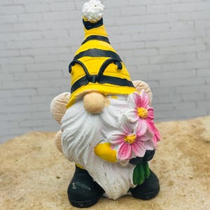 May include: A small gnome figurine with a yellow and black striped hat, a white beard, and a bouquet of pink flowers. The gnome is wearing black boots and has a white pointed hat.
