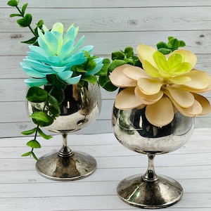 以下が含まれることがあります： ターコイズブルーとイエローの花の生け花が飾られた、銀色の金属製のベースが2つ。ターコイズブルーの花は多肉植物、イエローの花は紙の花です。