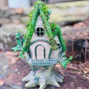 Puede incluir: Una caprichosa figura de casa de hadas. La casa en miniatura presenta una fachada de color claro con una pequeña puerta arqueada y una ventana redonda. El tejado está cubierto de musgo verde, y la casa está sobre una base de tronco de árbol.