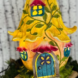 May include: A whimsical fairy house sculpture in the shape of a flower. The house features a yellow petal roof, orange walls, and a teal door with a purple flower design. Windows are trimmed in teal and topped with red flower petals.