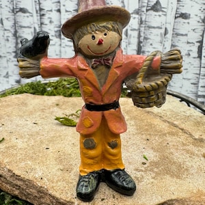 Könnte beinhalten: Eine skurrile Vogelscheuche-Figur mit einem braunen Hut, einer roten Jacke, einer gelben Hose und einem schwarzen Vogel, der auf ihrem Arm sitzt. Die Vogelscheuche hält einen Korb in der anderen Hand.