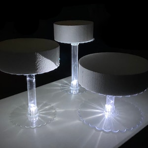 May include: Three illuminated cake stands with round, textured, gray platforms. The clear acrylic stems and bases have scalloped edges, and each stand has a bright light at the base. The background is dark.