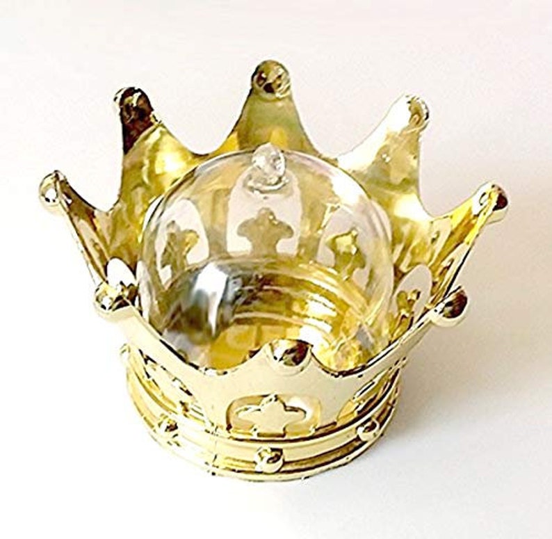 Puede incluir: Un recipiente dorado en forma de corona con una c&uacute;pula de cristal transparente. La corona tiene detalles ornamentados y un acabado brillante. La c&uacute;pula de cristal tiene un peque&ntilde;o pomo en la parte superior y est&aacute; dise&ntilde;ada para encajar en la corona.