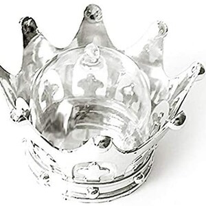 Puede incluir: Un recipiente plateado en forma de corona con una c&uacute;pula de cristal transparente. La corona tiene tres puntas prominentes y detalles decorativos. La c&uacute;pula de cristal se asienta en la base de la corona, creando una presentaci&oacute;n &uacute;nica.