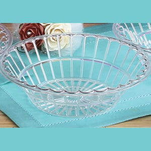 May include: Clear plastic oval-shaped basket with a lattice design. The basket has a scalloped edge and is sitting on a light blue surface. The background includes white and burgundy roses.