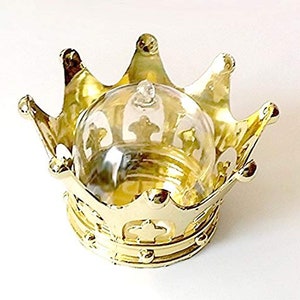 May include: A gold-colored crown-shaped container with a clear glass dome. The crown has ornate details and a shiny finish. The glass dome has a small knob on top and is designed to fit inside the crown.