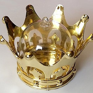 May include: A gold-colored crown-shaped decorative item with a clear, dome-shaped top. The crown features ornate details and a shiny finish, suitable for display or as a decorative accent. The crown has a base with small, round embellishments.