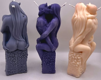 May include: Three sculpted candles depicting couples in an embrace. The candles are made of a smooth, waxy material and are painted in shades of purple and beige. The candles are displayed on a white surface.