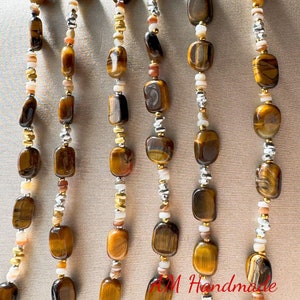 May include: A multi-strand necklace featuring brown tiger's eye beads, white and gold beads, and silver beads. The necklace is hanging against a light brown background.