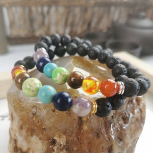 May include: Two bracelets with black lava rock beads and colourful gemstone beads. The bracelets are stacked on a light brown stone.