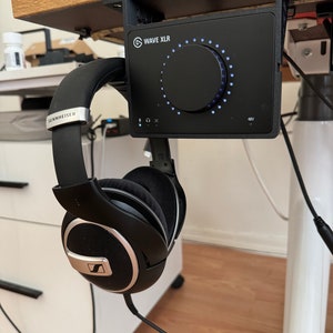 May include: A black Wave XLR audio interface with a large knob and a 48V indicator light. The interface is mounted under a desk with a black Sennheiser headphone hanging below.