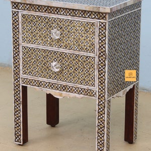 May include: A small, two-drawer side table with intricate inlaid mother-of-pearl and wood designs in shades of brown, white, and gold. The table has four legs and features crystal knobs on the drawers.