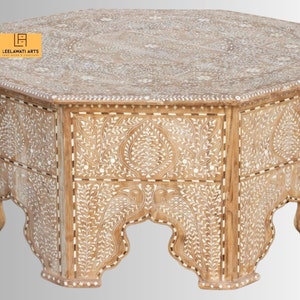 May include: A light brown, hexagonal coffee table with intricate white floral inlay designs. The table features a decorative edge and arched legs. The brand name "LEELAWATI ARTS" is visible on the top.