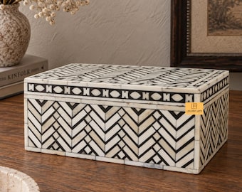 May include: A rectangular storage box featuring a black and white geometric herringbone pattern. The lid and sides are decorated with inlaid segments, and a horizontal band with an oval and bar motif encircles the top edge. A small orange logo reads LEELAWATI ARTS.
