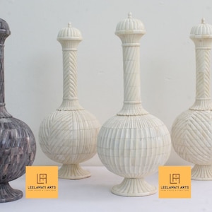 Peut inclure: Quatre vases décoratifs avec des sculptures complexes. Les vases sont en os blanc et gris et ont un corps rond et un long col fin. Les vases sont disposés en rangée sur une surface blanche. Le texte "LEELAWATI ARTS" est visible dans le coin inférieur droit de l'image.