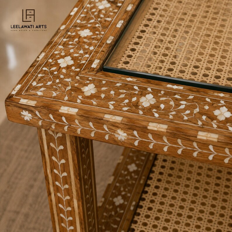 Pode incluir: Uma mesa lateral de madeira com intrincados padr&otilde;es de incrusta&ccedil;&atilde;o floral branca na moldura, uma prateleira de palhinha tran&ccedil;ada e um tampo de vidro. O log&oacute;tipo da Leelawati Arts &eacute; vis&iacute;vel, destacando o trabalho artesanal e a textura.