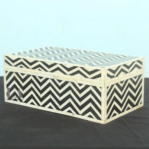 May include: Rectangular decorative box with a black and white chevron pattern. The box is made of a light-colored material, possibly wood or bone, with a contrasting black zigzag design. The box has a lid and is likely used for storage.