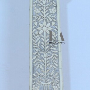 May include: Ivory-colored, carved pillar with floral and leaf patterns. The pillar has a square column with detailed carvings of flowers and foliage. The base and top are also intricately designed, showcasing a classic, elegant aesthetic.
