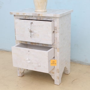 May include: White nightstand with two drawers, made of inlaid mother of pearl. The top drawer has a keyhole and the bottom drawer has a crystal knob. The nightstand has a small label on the bottom right side that says "Leelavati Arts".