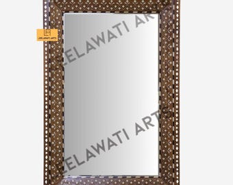 Può includere: Specchio rettangolare con cornice in legno marrone e intricati motivi intarsiati. La cornice presenta un design geometrico ripetuto con dettagli bianchi e marroni. Lo specchio ha una superficie riflettente e un piccolo logo.