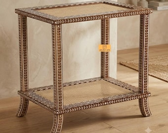 Pode incluir: Uma mesa lateral retangular com estrutura castanha, incrustações florais brancas e dois níveis de prateleiras de cana entrançada. A mesa assenta em quatro pernas curvas e inclui uma pequena etiqueta quadrada laranja com o texto Leelawati Arts.