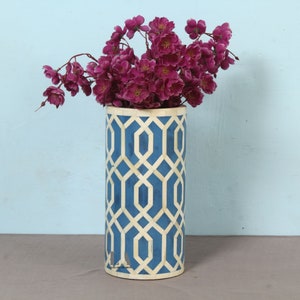 May include: A cylindrical vase with a cream and blue geometric pattern, filled with deep magenta flowers. The vase has a cream rim and base. The flowers are in full bloom. The vase is a decorative item.