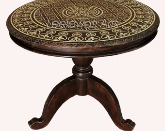 Op de afbeelding: Donkerbruine, ronde bijzettafel met ingewikkelde, lichtgekleurde inlegpatronen op het tafelblad. De tafel heeft een voetstuk met drie gebogen poten. De tekst "Leelawati Arts" is zichtbaar op het tafelblad.