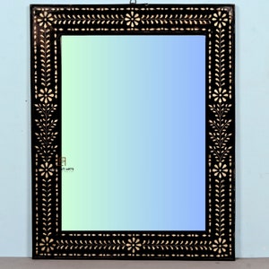Puede incluir: Espejo rectangular con marco negro y diseño floral. El marco presenta un intrincado patrón de detalles florales y de hojas blancas. La superficie del espejo refleja un degradado de tonos azules y verdes, creando un acento decorativo de pared.