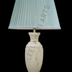 Può includere: Lampada da tavolo decorativa con paralume plissettato azzurro e base color crema. La base presenta un design a mosaico e una forma slanciata ed elegante. La lampada è ambientata su uno sfondo nero.
