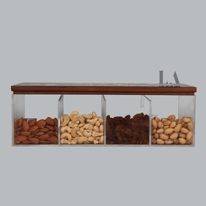 May include: A wooden tray with four clear acrylic containers filled with different types of nuts. The containers are filled with almonds, cashews, raisins, and pistachios.