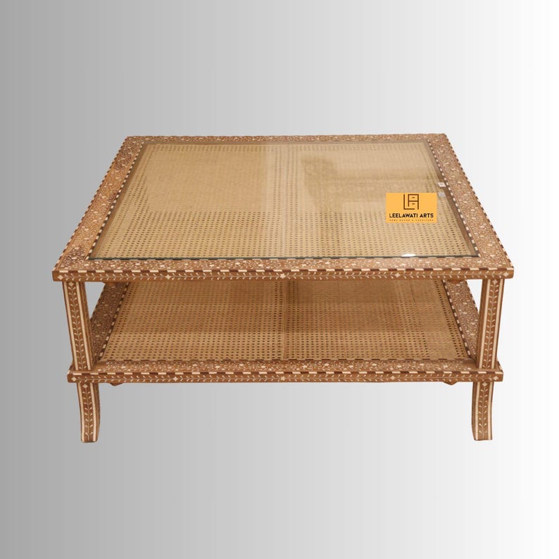 Pode incluir: Uma mesa de centro quadrada com tampo de vidro e prateleira inferior, ambas com superf&iacute;cies de palhinha tran&ccedil;ada. A estrutura de madeira &eacute; decorada com intrincados padr&otilde;es florais brancos incrustados. Uma etiqueta amarela com o texto LEELAWATI ARTS &eacute; vis&iacute;vel.