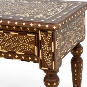May include: A dark brown wooden side table featuring intricate cream-colored floral and geometric inlay patterns across the top, drawer front, and legs. The table has a rectangular top, a single drawer, and four turned legs with inlaid details.