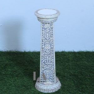May include: A tall, off-white decorative pedestal with intricate floral carvings. The pedestal features a round top and base, with a long, rectangular column. The column is adorned with detailed floral patterns and a vase design. The pedestal is suitable for displaying sculptures or plants.