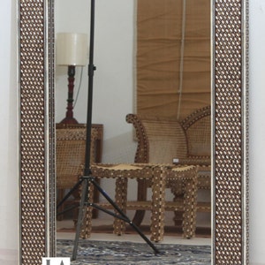 May include: Rectangular mirror with a decorative frame. The frame features a repeating pattern of small, light-colored shapes set against a dark brown background. The mirror reflects a room with furniture and a lamp.