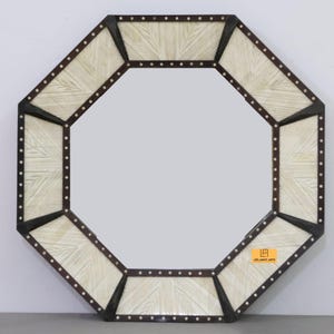 Könnte beinhalten: Achteckiger Spiegel mit dunkelbraunem Rahmen und dekorativer weiß-cremefarbener Intarsie. Der Rahmen ist mit kleinen, gleichmäßig verteilten silberfarbenen Nieten versehen. Das Design des Spiegels ist geometrisch und modern.