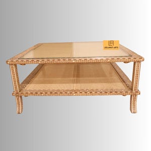 Pode incluir: Uma mesa de centro quadrada com tampo de vidro e prateleira inferior com detalhes em palhinha tran&ccedil;ada. A estrutura de madeira &eacute; decorada com padr&otilde;es de incrusta&ccedil;&otilde;es florais brancas. Uma etiqueta laranja exibe o texto LEELAWATI ARTS.