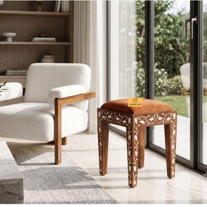 Puede incluir: Un taburete de madera con un asiento cuadrado tapizado en color naranja quemado y cuatro patas con patrones florales blancos incrustados. El taburete tiene una pequeña etiqueta rectangular con el texto Leelamati Arts en el cojín.
