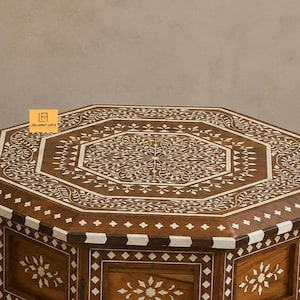 May include: An octagonal wooden table featuring intricate white floral and geometric inlay patterns across the top and sides. The surface displays a central medallion with leaf motifs, bordered by a repeating diamond and chevron inlay design.