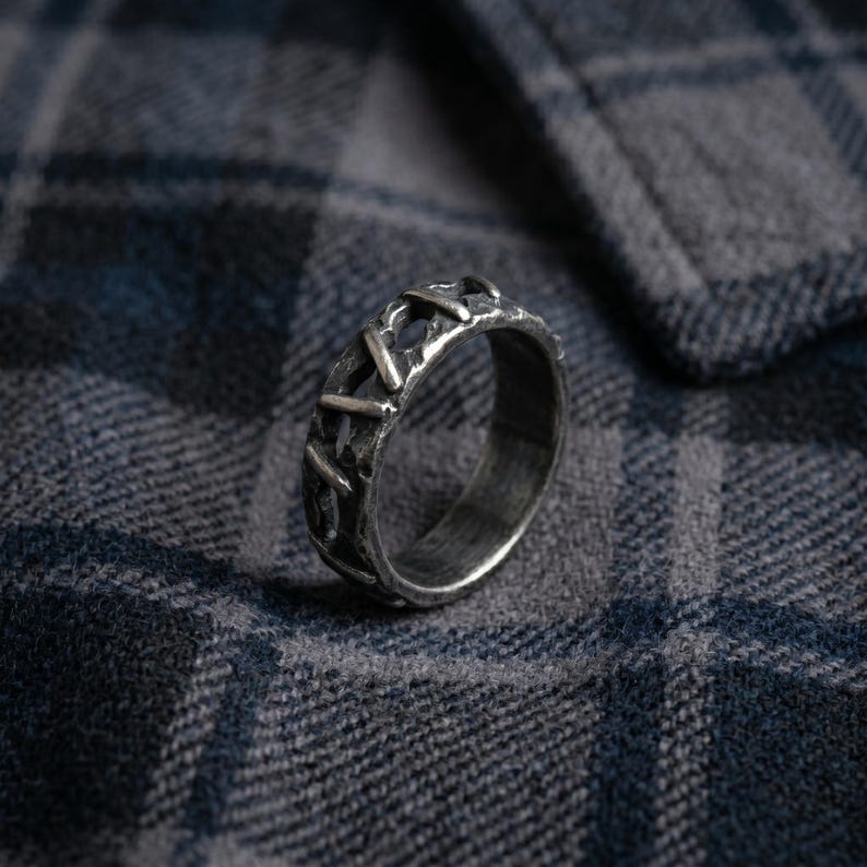 Op de afbeelding: Een zilverkleurige metalen ring met een donkere, geoxideerde afwerking en een getextureerd, ruw oppervlak met een reeks verhoogde, diagonale kruisarceringen rond de gehele buitenband, wat een uitgesproken, verweerde en industri&euml;le uitstraling geeft.