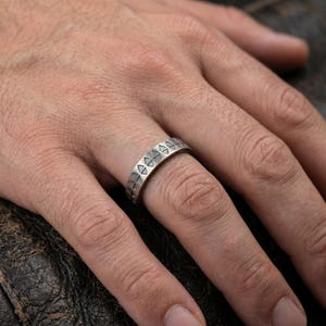May include: A silver-toned metal ring featuring a repeating geometric pattern of small triangles etched around the entire band. The ring has a matte, oxidised finish that highlights the engraved triangular shapes across its surface.