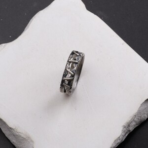 Op de afbeelding: Een zilverkleurige metalen ring met een getextureerde, geoxideerde afwerking en een herhalend patroon van verhoogde, diagonale, balkvormige elementen die lijken op steken rondom de gehele buitenomtrek van de band.