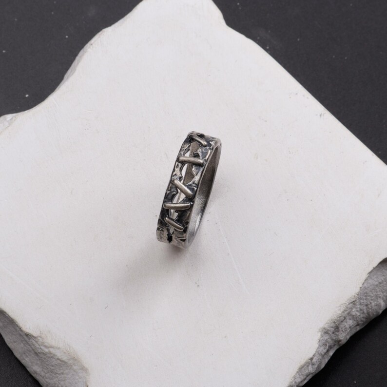 Op de afbeelding: Een zilverkleurige metalen ring met een getextureerde, geoxideerde afwerking en een herhalend patroon van verhoogde, diagonale, balkvormige elementen die lijken op steken rondom de gehele buitenomtrek van de band.