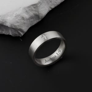 May include: A silver ring with a cross engraved on the top and the text "Joshua 1:9" on the side. The ring is made of a brushed metal and sits on a dark surface.