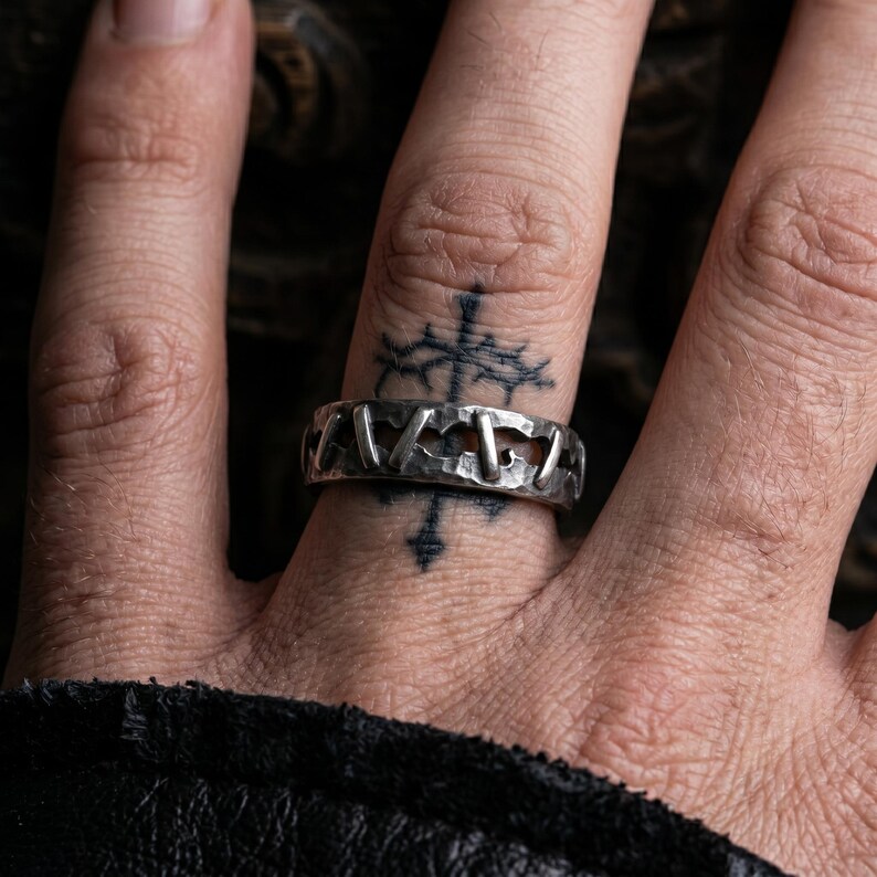 Op de afbeelding: Een zilverkleurige metalen ring met een getextureerd, gehamerd oppervlak en een reeks gelijkmatig verdeelde, verticale, staafachtige inkepingen rondom de band. De ring heeft een donkere, geoxideerde afwerking in de uitsparingen voor contrast.