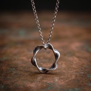 May include: A silver-toned necklace featuring a circular pendant with a twisted, undulating edge. The pendant hangs from a simple round-link chain. The metal has a dark, oxidized finish with highlights on the curved surfaces of the ring.